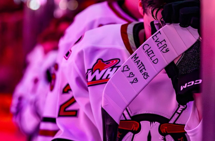 Dwayne Jean Jr. stands on the bench with his head down. On his stick are the words 