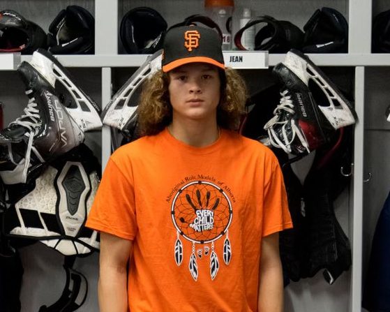 Dwayne Jean Jr. stands in front of a locker room stall. He has a black hat and an Orange Shirt Society shirt on.